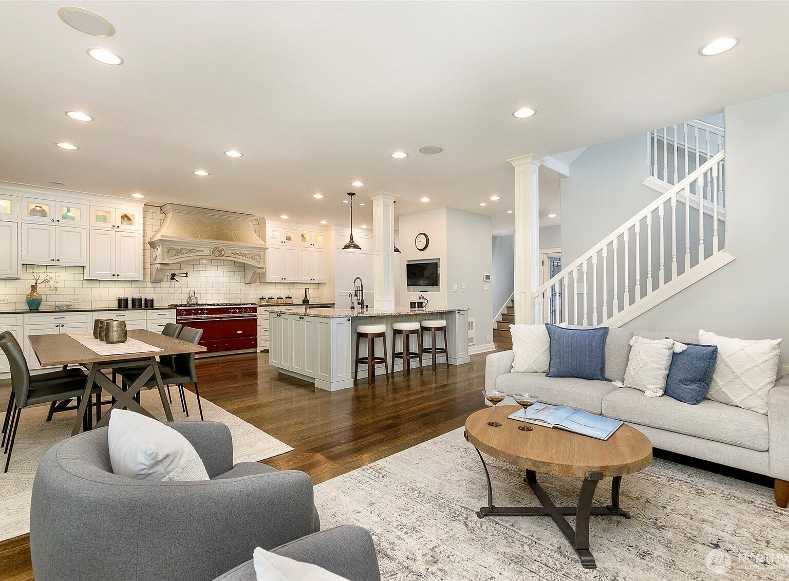 Open great room and chef's kitchen with a red Lacanche range under a custom vent hood, oversized island with seating, recessed lighting throughout, and the staircase rising at the right